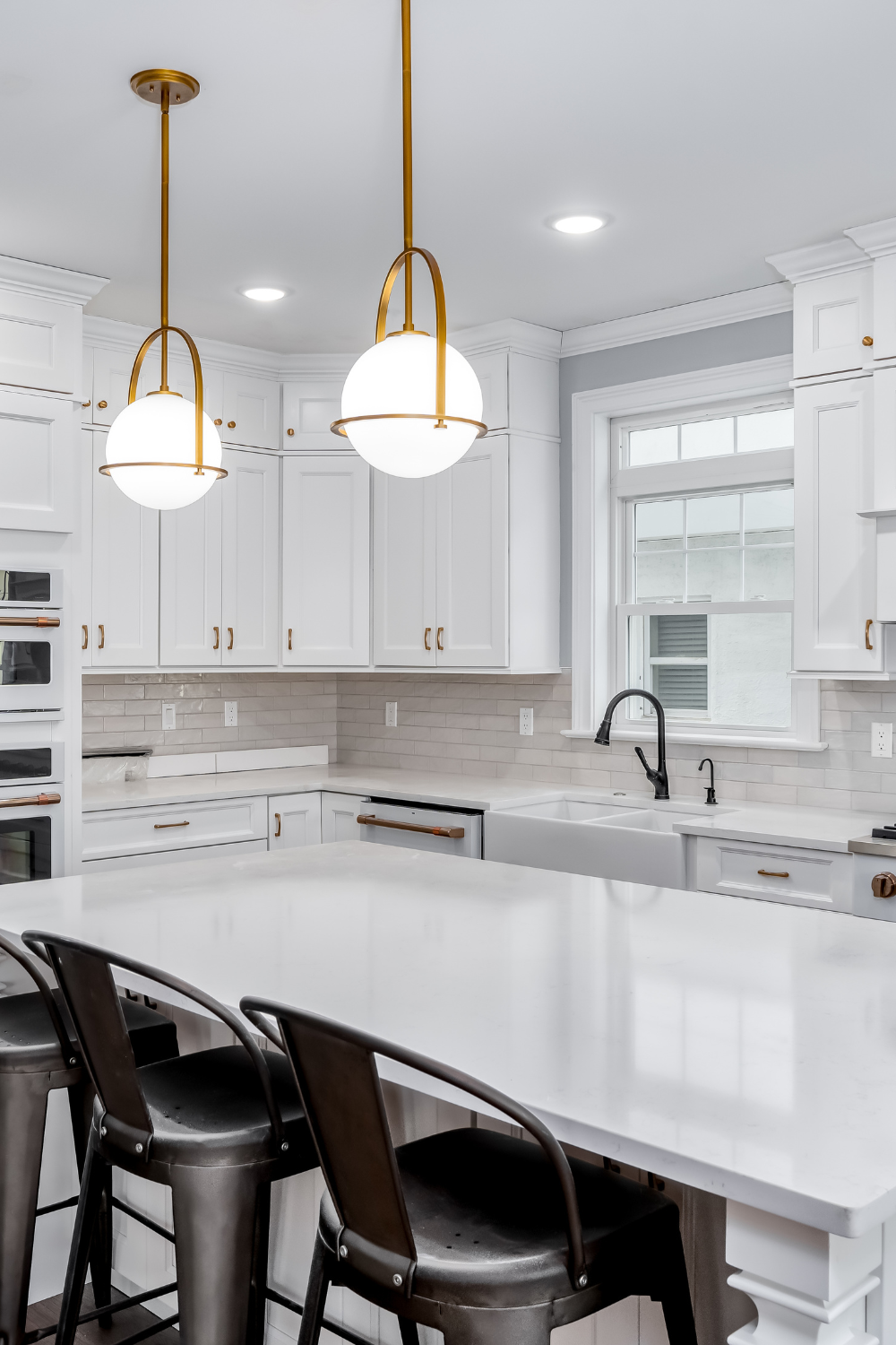 A contemporary kitchen with white cabinets, white tile backsplash, black chairs, and gold lamp fixtures.
